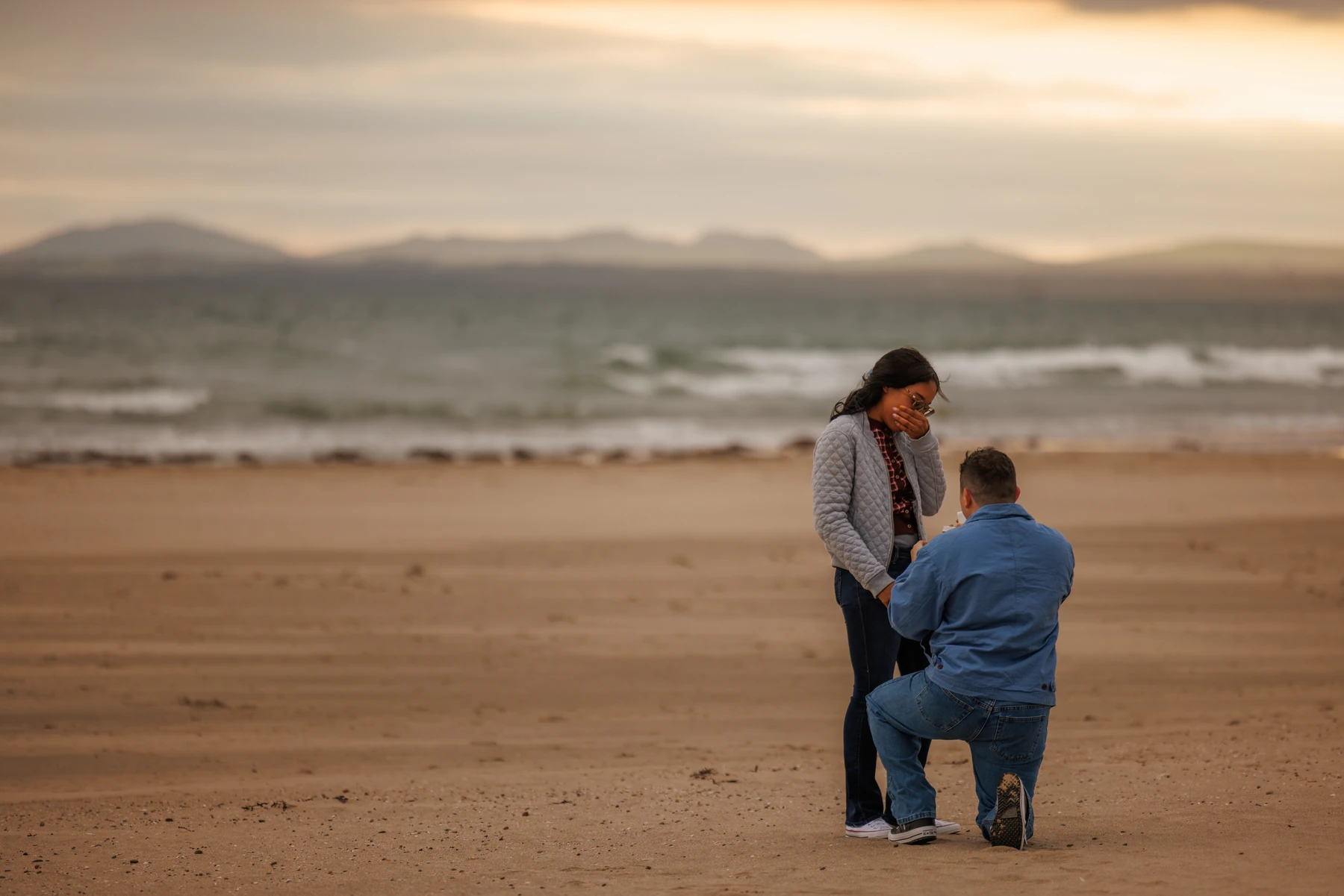 A surprise proposal on the beach at sunset, with the sea and distant hills behind the couple
