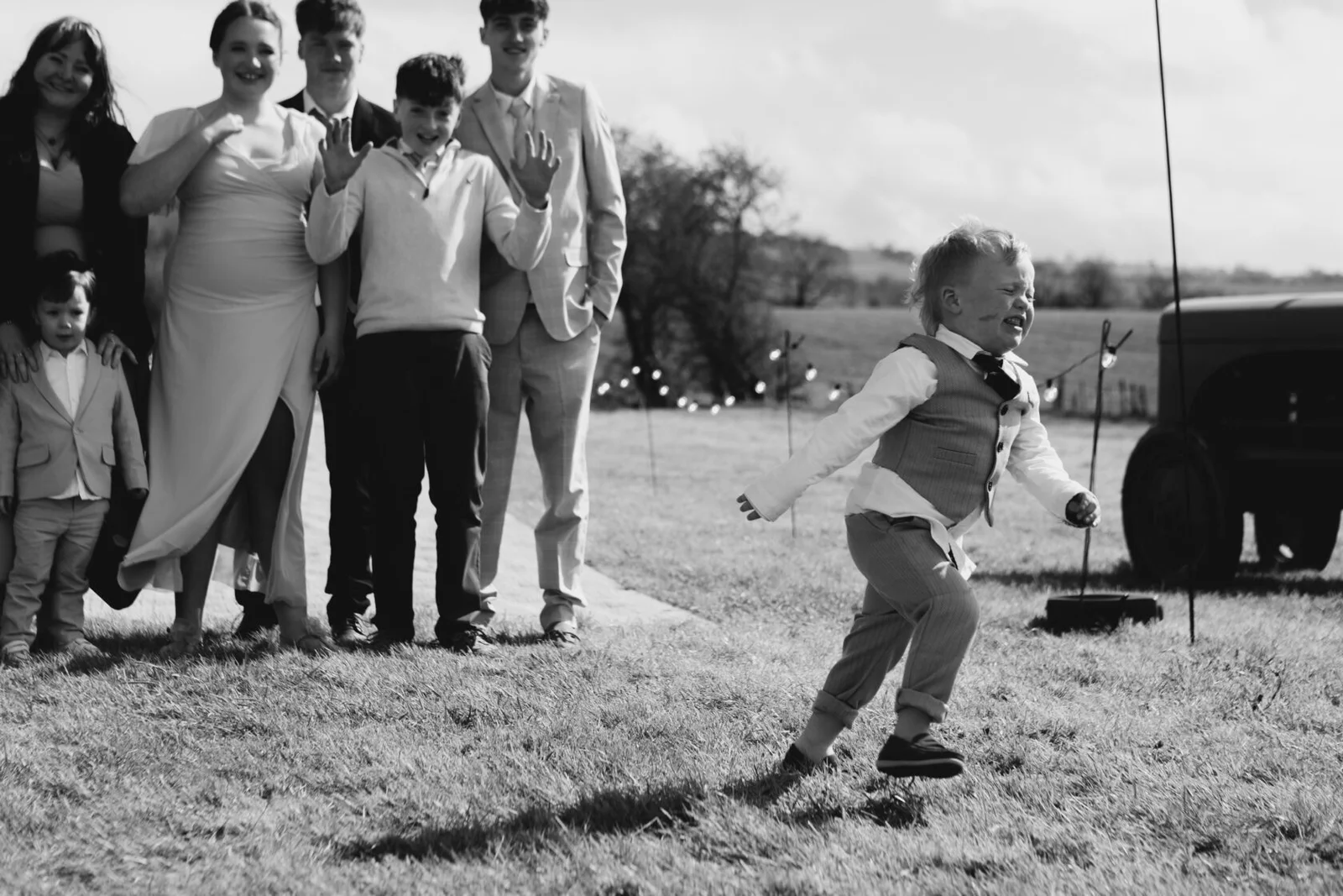 A child running across the grass in front of wedding guests, in black and white